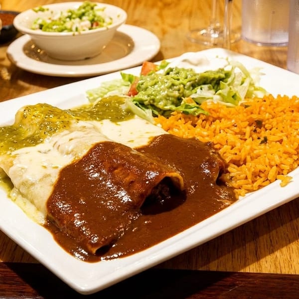 A plate of enchiladas with green and mole sauce, rice, and salad, with chips and salsa in the background.