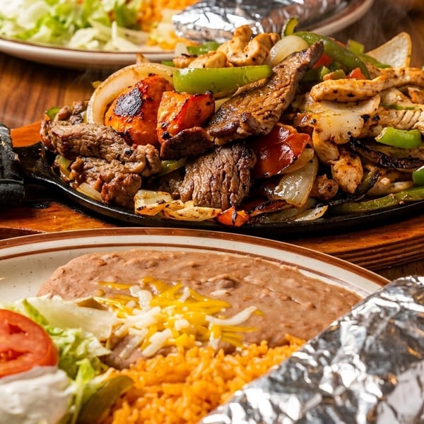 Sizzling fajitas with steak, chicken, and vegetables, alongside a plate of rice, beans, and salad.