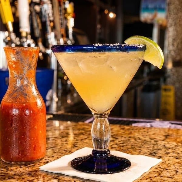 Margarita with a lime wedge and salt rim, next to a carafe of red salsa on a bar.