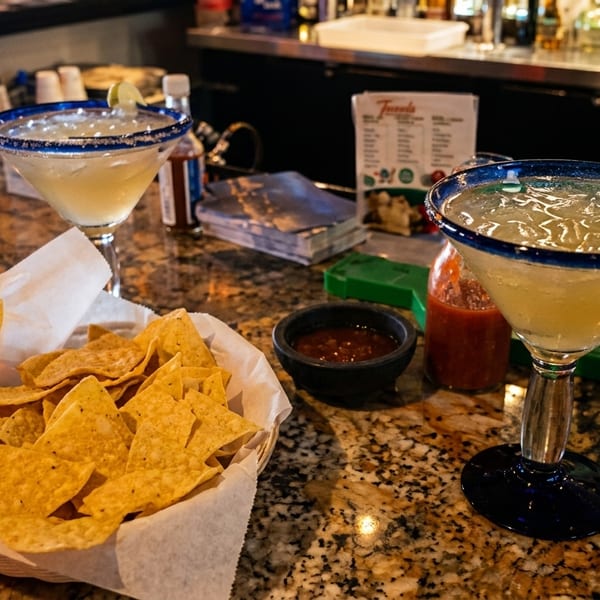 Two margaritas, a basket of tortilla chips, and salsa on a granite bar top.